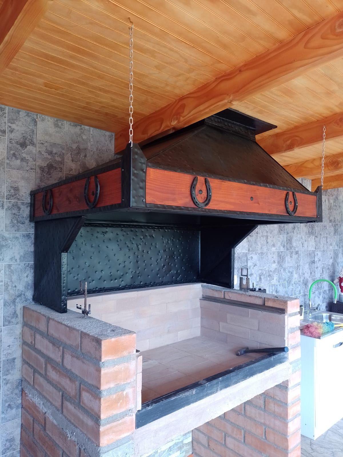 Campana de plancha martillada negra con banda de madera y herraduras, instalada sobre un asador de ladrillo en un quincho